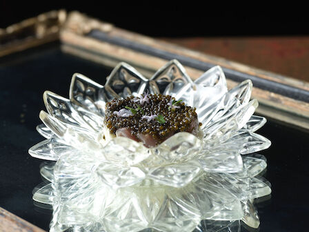 A decorative glass dish shaped like a flower holds creamy dessert with a topping that appears to be herbs or caviar on a reflective surface.