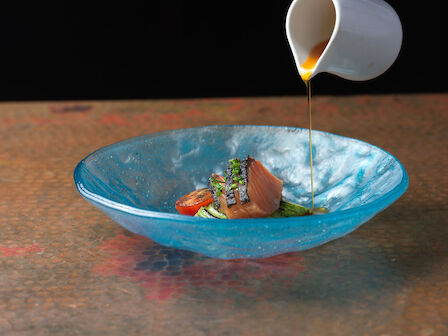A hand pours sauce from a small jug onto a dish of salmon and vegetables in a blue bowl on a table.