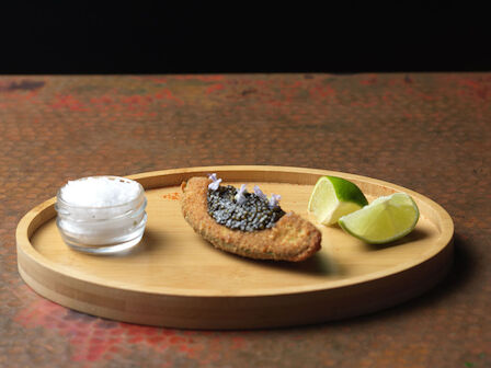 A wooden tray with caviar on bread, lime wedges, and a small jar of salt on a dark background.