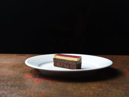 A slice of layered dessert with a dark base, light middle layer, and red top sits on a white plate against a dark background.
