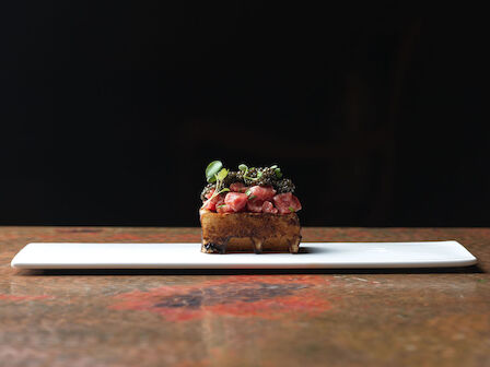 A small, elegantly plated dish sits on a rectangular platter with a dark background, featuring meat and garnishes.