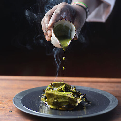 A person pours green sauce over a steaming dish on a dark plate, creating a visually striking culinary scene.