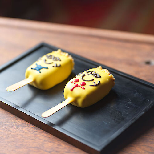 Two yellow ice cream bars decorated with cartoon faces and colored details, set on a black rectangular tray on a wooden surface.