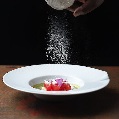 A hand sprinkles a fine powder over a dish with garnish, using a sieve, set against a dark background.