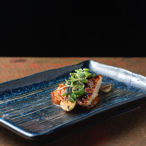 A gourmet dish is presented on a blue rectangular plate, garnished with herbs and vegetables, set against a dark background.