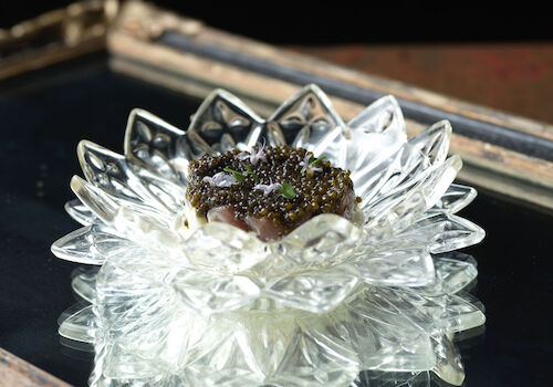 A decorative glass dish with an ornate design holds a scoop of creamy dessert topped with dark garnish, set on a reflective surface.