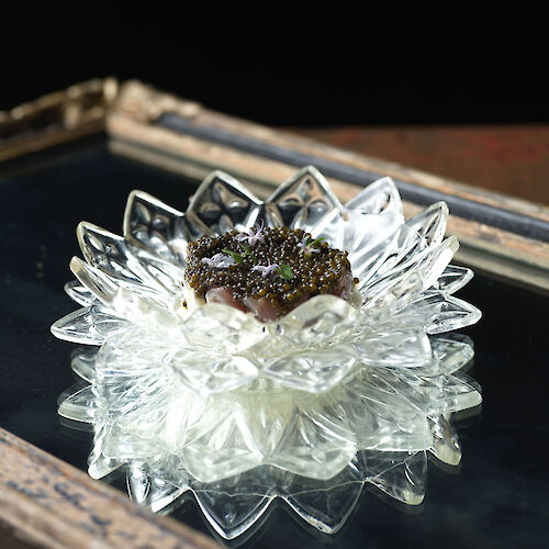 A decorative glass dish with an ornate design holds a scoop of creamy dessert topped with dark garnish, set on a reflective surface.