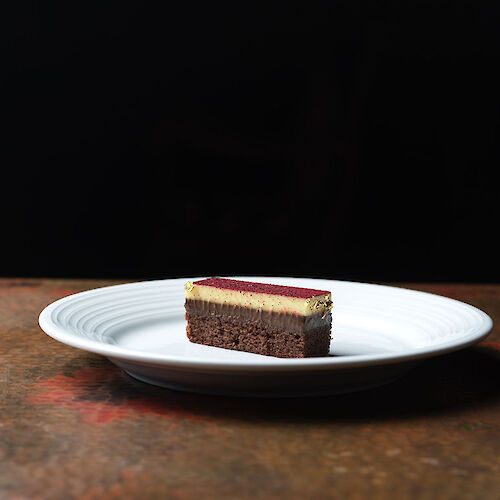 A slice of layered cake with a chocolate base and cream topping sits on a white plate against a dark background.