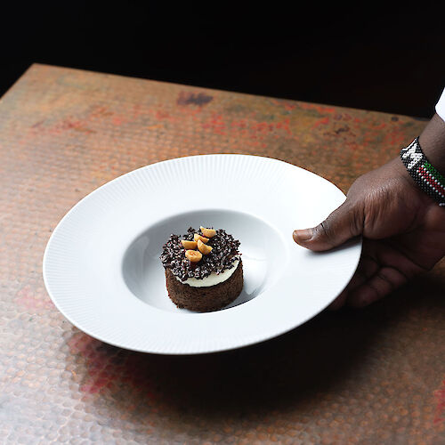A hand is holding a white plate with a chocolate dessert garnished with nuts on a textured brown surface.