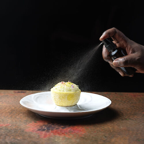 A hand spraying a mist onto a yellow dessert on a white plate against a dark background.
