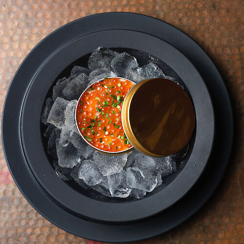 A tin of orange fish roe garnished with herbs, placed on ice in a dark bowl on a textured surface.
