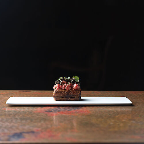 A small gourmet dish with a dark background on a long white plate, featuring fresh ingredients and garnishes.