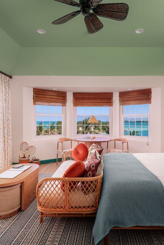 A serene bedroom overlooks the ocean, featuring a cozy bed, seating area, and ceiling fan, with large windows revealing a beautiful seaside view.
