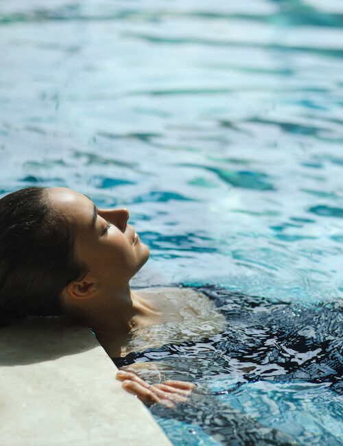 A person is relaxing in a swimming pool, leaning back against the edge, enjoying the water and sunlight.