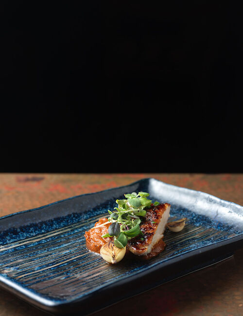 A piece of grilled fish garnished with green herbs and seeds, served on a rectangular blue plate against a dark background.
