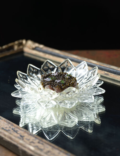 A decorative glass dish with a reflective base holds an intricately designed food item, possibly garnished, set against a dark backdrop.