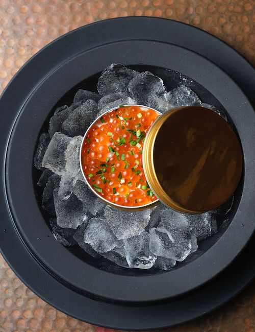 A tin of red caviar garnished with herbs sits on ice in a dark dish on a textured surface.
