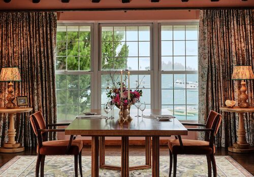 A cozy dining room features a wooden table, chairs, floral arrangement, large windows with a lake view, and a woven chandelier.