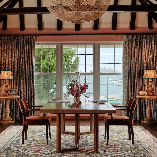 A cozy dining room features a wooden table, chairs, floral arrangement, large windows with a lake view, and a woven chandelier.