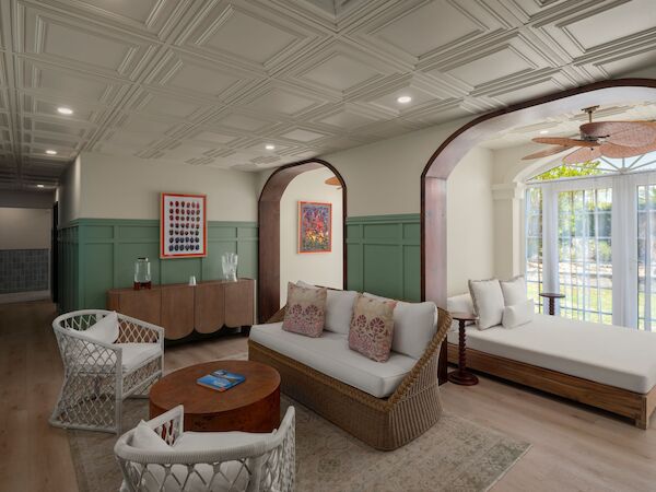 A cozy living room with wicker furniture, arched doorways, decorative ceiling, and art on walls, featuring a view of greenery outside.