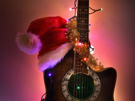 A guitar wearing a Santa hat with festive lights and tinsel draped around it, set against a warm, colorful background.
